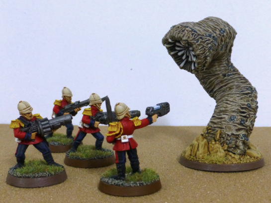 Four soldiers in red jackets aiming guns at a large worm that burst from the desert floor