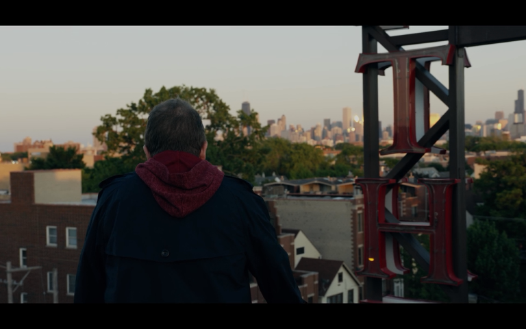 Patton Oswalt is seen from the back in a hoodie and jacket, looking over the city of Chicago during a sunset that is starting off beautifully.